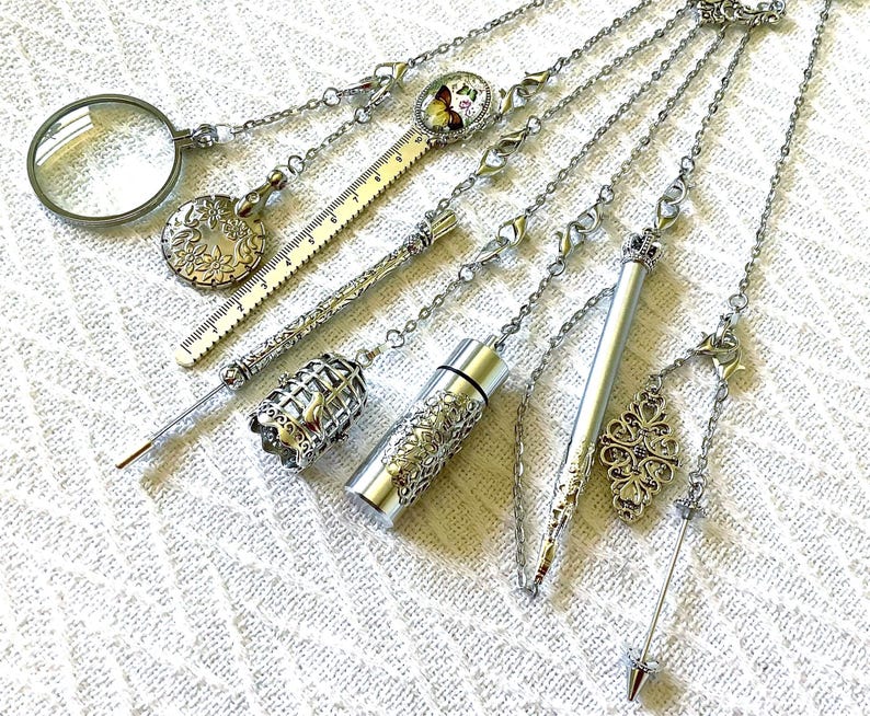 May include: A collection of silver-toned metal accessories attached to chains, including a magnifying glass, a floral embossed disc, a ruler, a birdcage charm, a cylindrical container, a slender pen, and a pointed tool with a decorative filigree charm.