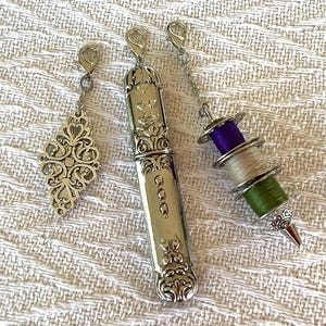 May include: Three silver sewing accessories: a decorative charm, a needle case, and a thread spool charm. The needle case has ornate floral detailing and the thread spool charm has purple, green, and beige thread.