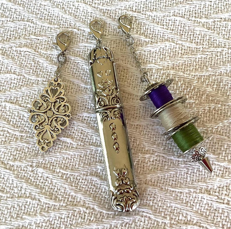 May include: Three silver sewing accessories: a decorative charm, a needle case, and a thread spool charm. The needle case has ornate floral detailing and the thread spool charm has purple, green, and beige thread.