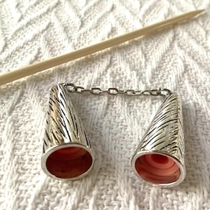 May include: Silver-toned metal knitting needle protectors connected by a chain. The cone-shaped protectors have a textured design and a pink interior. A wooden knitting needle rests diagonally in the background.