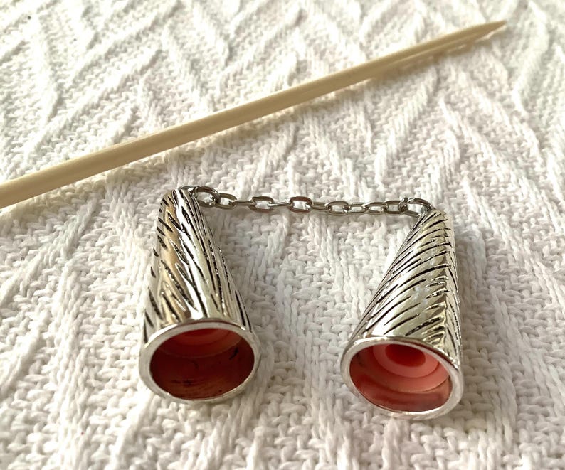 May include: Silver-toned metal knitting needle protectors connected by a chain. The cone-shaped protectors have a textured design and a pink interior. A wooden knitting needle rests diagonally in the background.