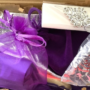May include: A cardboard box containing a purple velvet pouch, a purple organza drawstring bag, a silver metallic fabric pouch, and a white rectangular card featuring an ornate silver filigree metal embellishment with a central floral motif.