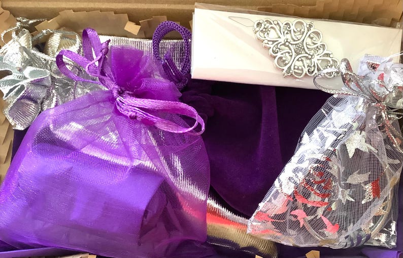 May include: A cardboard box containing a purple velvet pouch, a purple organza drawstring bag, a silver metallic fabric pouch, and a white rectangular card featuring an ornate silver filigree metal embellishment with a central floral motif.