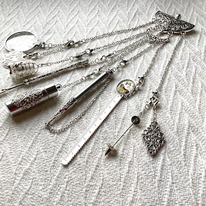 May include: A silver-toned multi-charm accessory featuring a decorative top piece with dangling chains attached to a magnifying glass, a birdcage, a metal tube, a ruler with a butterfly image, a pointed needle tool, and a filigree diamond shape.