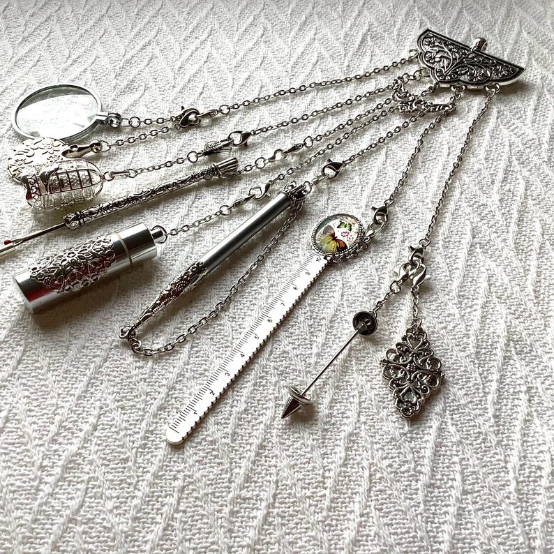May include: A silver-toned multi-charm accessory featuring a decorative top piece with dangling chains attached to a magnifying glass, a birdcage, a metal tube, a ruler with a butterfly image, a pointed needle tool, and a filigree diamond shape.