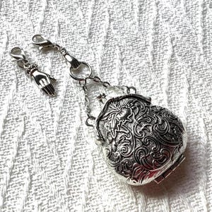 May include: A silver-toned metal charm shaped like a miniature coin purse with an intricate embossed floral pattern. The charm features a hinged clasp closure and is attached to a lobster claw hook, accompanied by a separate small hand-shaped charm.