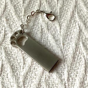 May include: A silver USB flash drive with a paw print design at the top, connected to a chain and clasp. The drive is rectangular and has a smooth, metallic finish. The chain and clasp are also silver.
