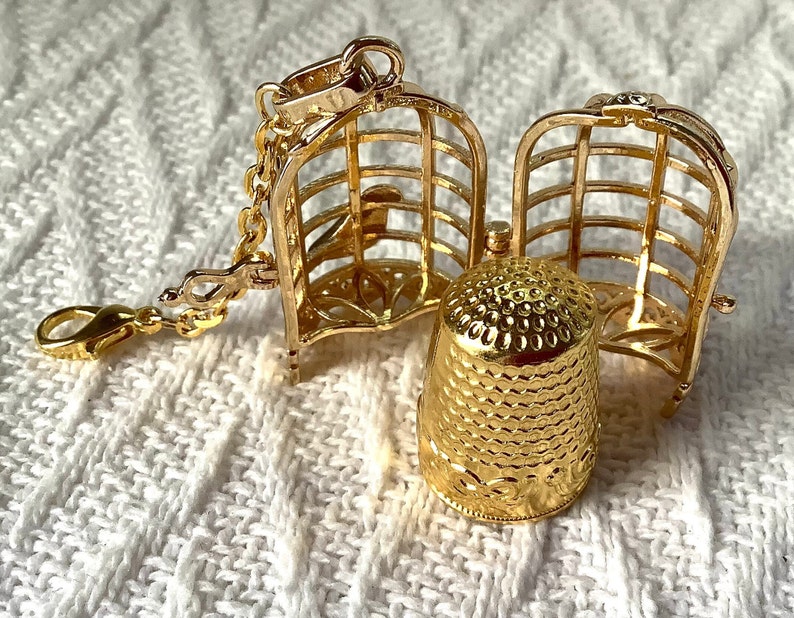May include: A gold-toned metal thimble charm with a cage design that opens to reveal the thimble inside. The charm has a small chain attached to it.