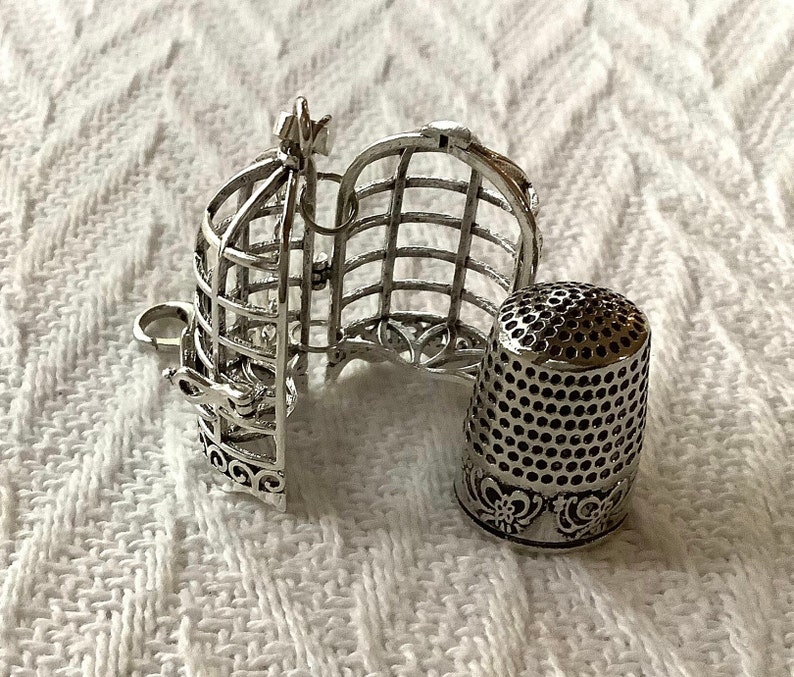 May include: A silver metal thimble shaped like a birdcage with the door open. The thimble is sitting on a white knitted surface.