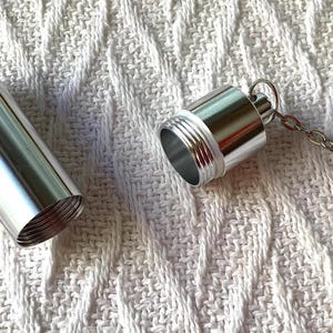 May include: A cylindrical silver metal pill container with a screw-top lid. The lid is detached from the main body and features a short silver chain with a lobster claw clasp attached to the top, allowing it to be used as a keychain accessory.