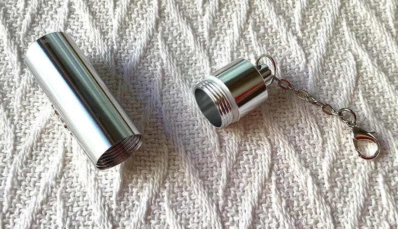 May include: A cylindrical silver metal pill container with a screw-top lid. The lid is detached from the main body and features a short silver chain with a lobster claw clasp attached to the top, allowing it to be used as a keychain accessory.
