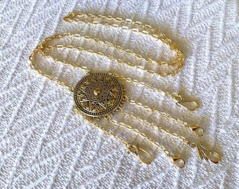 May include: Gold-toned necklace with a circular pendant featuring an intricate sunburst design. The necklace has multiple strands with lobster claw clasps. The jewelry is displayed on a white, textured surface.