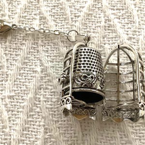 May include: A silver-toned metal charm featuring a hinged cage that opens to reveal a thimble inside. The cage has vertical bars and decorative scrollwork at the base, while the thimble has a textured dimpled surface and a bow detail.