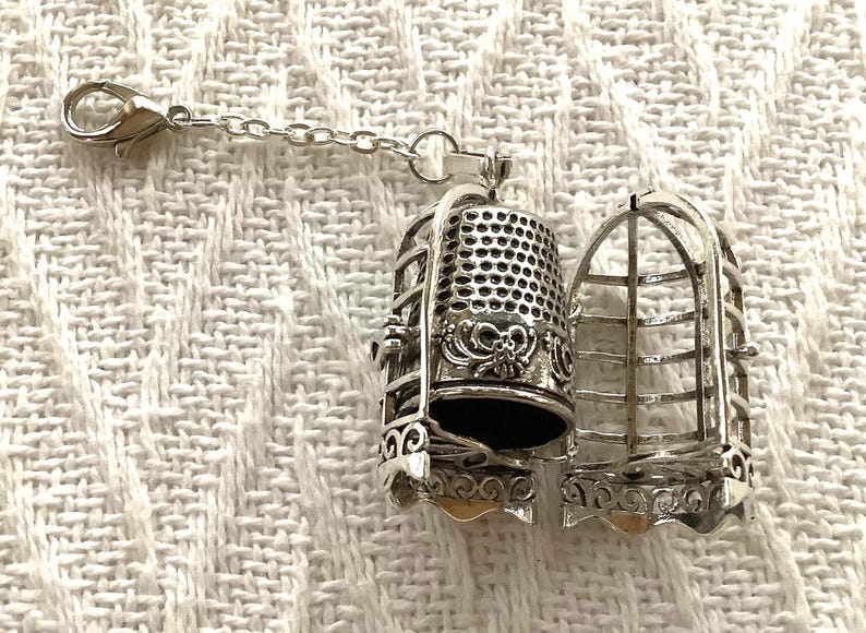 May include: A silver-toned metal charm featuring a hinged cage that opens to reveal a thimble inside. The cage has vertical bars and decorative scrollwork at the base, while the thimble has a textured dimpled surface and a bow detail.