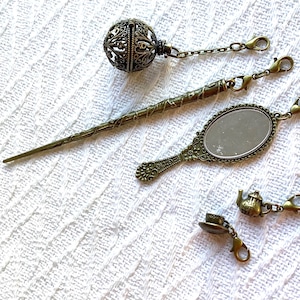 May include: A set of antique bronze-toned metal charms and accessories including a filigree sphere, a long textured hair stick, an oval hand mirror, a miniature teapot, and a tiny teacup, each attached to a small lobster claw clasp.
