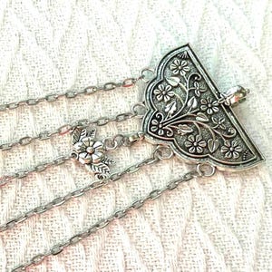 May include: Silver-toned, multi-strand body jewelry. The piece features a triangular, floral-patterned centerpiece and delicate chain links. Additional floral accents are incorporated along the chains, creating an elegant, bohemian aesthetic.