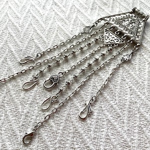 May include: Silver-toned metal pendant with intricate geometric and floral designs. The pendant features a triangular top section with dangling chains of varying lengths, some with decorative beads and clasps. The piece is displayed on a white, textured surface.