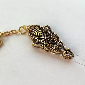 May include: A gold-colored, ornate charm with a decorative filigree design. The charm is attached to a small gold chain and a lobster clasp. A needle is attached to the charm.