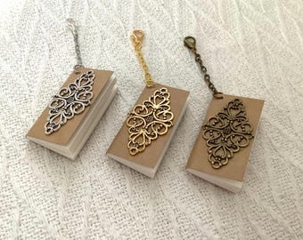 May include: Three miniature books with tan covers and white pages, each featuring a decorative metal filigree overlay on the front. The books are attached to silver, gold, and bronze-toned metal chains with lobster claw clasps.