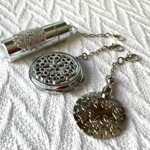 May include: Silver tea infuser set with a round, ornate tea strainer, a small, cylindrical tea container, and a decorative, round, floral-patterned charm. The set is connected by a silver chain.