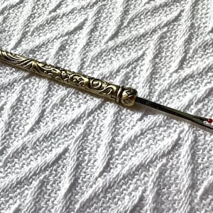 May include: A gold-toned metal seam ripper featuring an ornate, embossed floral pattern along the handle. The tool includes a thin, sharp blade with a small red safety ball at the tip and a separate, matching gold-toned metal protective cap.