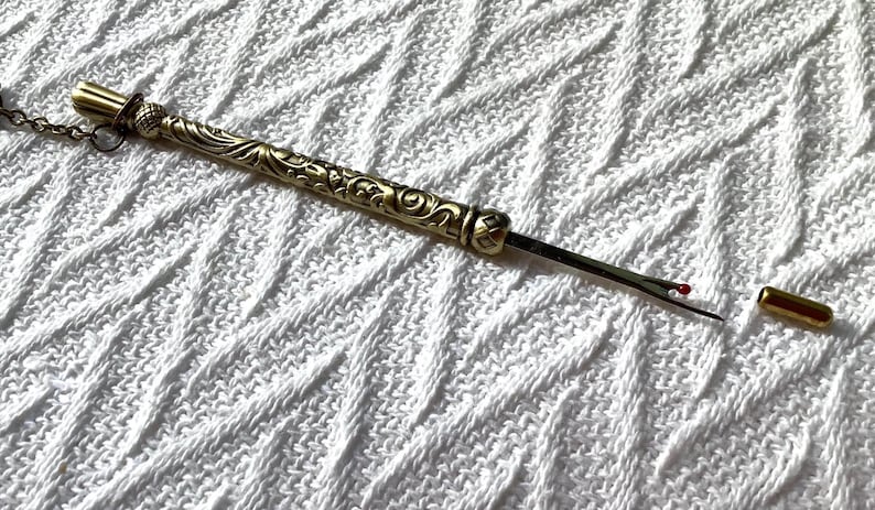 May include: A gold-toned metal seam ripper featuring an ornate, embossed floral pattern along the handle. The tool includes a thin, sharp blade with a small red safety ball at the tip and a separate, matching gold-toned metal protective cap.
