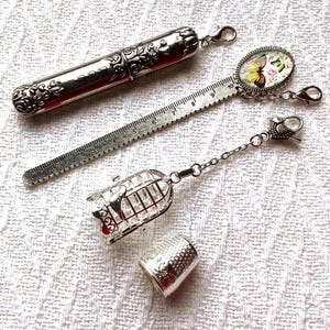 May include: A silver-toned needle case with ornate floral embossing, a metal ruler charm featuring a butterfly graphic, a birdcage charm with a small bird inside, and a silver-toned thimble with a textured honeycomb pattern.