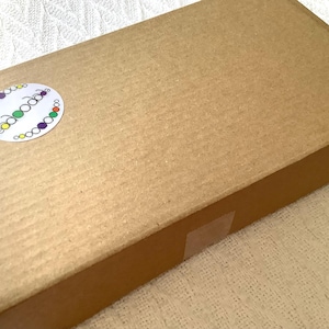 May include: A rectangular brown cardboard shipping box with a corrugated texture. A circular white sticker is affixed to the top left corner, featuring the text "beadoodle" in a lowercase font surrounded by a border of small, colorful circles.