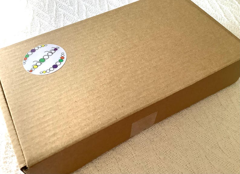 May include: A rectangular brown cardboard shipping box with a corrugated texture. A circular white sticker is affixed to the top left corner, featuring the text "beadoodle" in a lowercase font surrounded by a border of small, colorful circles.