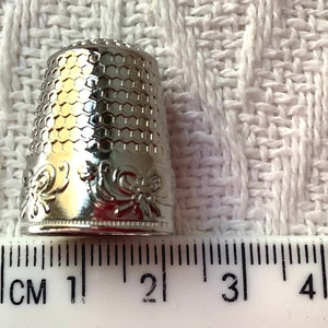 May include: Silver thimble with a hexagonal pattern and floral design. The thimble is 2 cm tall and is measured against a ruler.