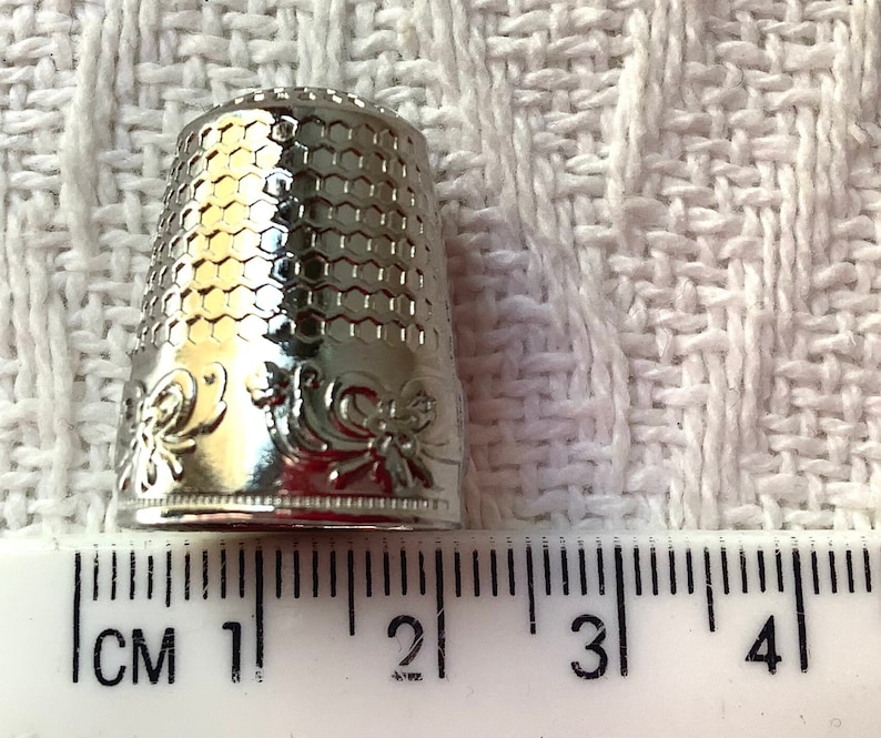 May include: Silver thimble with a hexagonal pattern and floral design. The thimble is 2 cm tall and is measured against a ruler.