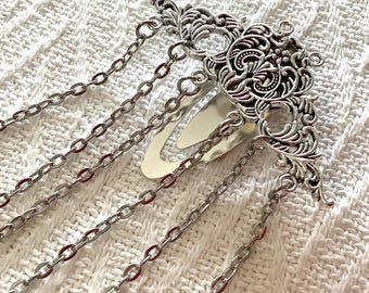 May include: A silver-toned hair clip featuring an ornate, filigree-style metal decorative piece attached to a snap-style barrette. Five delicate silver-coloured metal chains of varying lengths hang from the decorative top piece, creating a layered effect.