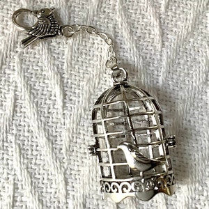 May include: A silver birdcage charm with a small bird inside. The cage has a detailed design and is attached to a silver chain with a bird-shaped clasp.