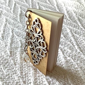 May include: A miniature book charm featuring a tan cover adorned with an intricate silver-toned filigree metal embellishment. The small book has multiple white pages and is attached to a silver-coloured metal chain with a lobster claw clasp.