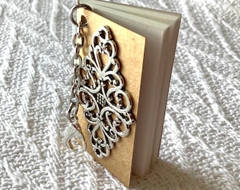 May include: A miniature book charm featuring a tan cover adorned with an intricate silver-toned filigree metal embellishment. The small book has multiple white pages and is attached to a silver-colored metal chain with a lobster claw clasp.
