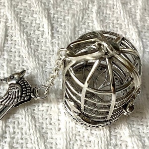 May include: Silver birdcage charm with a small bird attached to the side. The cage is detailed with intricate designs and has a small loop at the top for attaching to a necklace or bracelet.