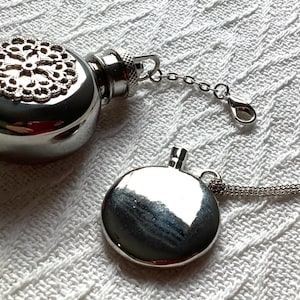 May include: A silver-toned metal flask pendant with a circular shape and a decorative filigree top piece. The flask features a short chain with a lobster clasp attached to the neck. A second, flat, circular silver-toned metal pendant hangs nearby.