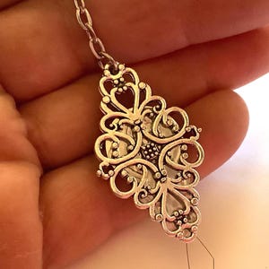May include: Silver-toned filigree pendant with an ornate, symmetrical design. The pendant is attached to a delicate chain and features a small, diamond-shaped charm at the bottom. The intricate details and openwork create a vintage aesthetic.