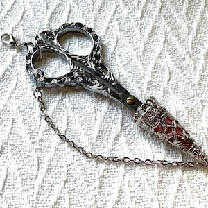 May include: A silver-toned metal scissor pendant featuring ornate floral detailing on the handles and a brass-colored screw. The pointed blades are encased in a decorative, filigree-style silver cone with a reddish-pink inlay and a thin chain.