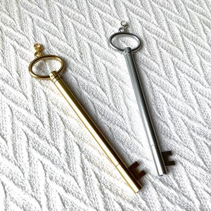 May include: Two key-shaped pens, one gold-toned and one silver-toned, lie on a white, textured surface. The pens have a ring at the top and a key-shaped design at the bottom. The gold pen has a small clasp attached to the ring.