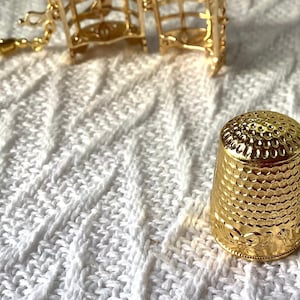 May include: A gold-colored thimble with a textured surface and a decorative design around the base. The thimble is sitting on a white knitted surface.