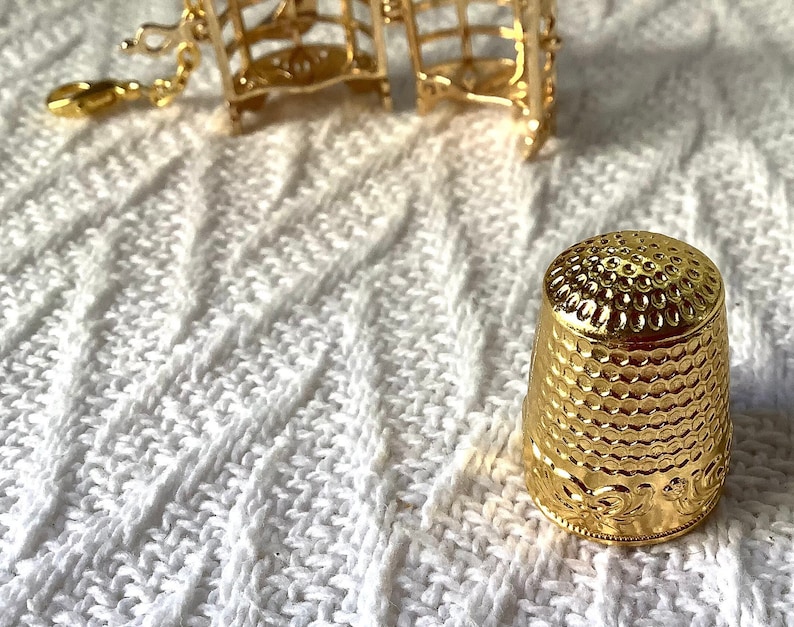 May include: A gold-colored thimble with a textured surface and a decorative design around the base. The thimble is sitting on a white knitted surface.