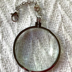 May include: A silver-toned magnifying glass with a clear lens and a decorative chain. The chain has a lobster clasp and a small, round, clear gem. The magnifying glass is round and has a silver frame.