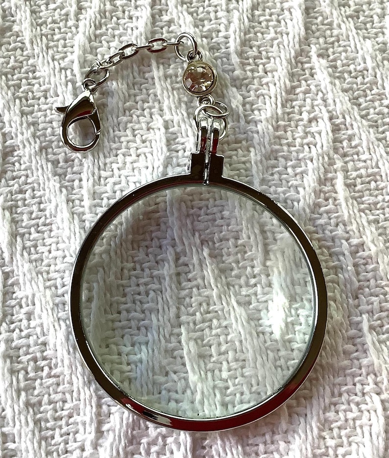 May include: A silver-toned magnifying glass with a clear lens and a decorative chain. The chain has a lobster clasp and a small, round, clear gem. The magnifying glass is round and has a silver frame.