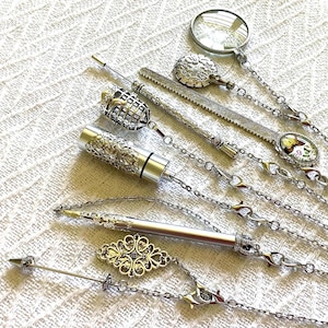 May include: A collection of silver-toned metal accessories including a magnifying glass, a ruler, a cylindrical container with filigree detail, a pen, and decorative charms, all connected by delicate silver chains with small clear crystal accents.