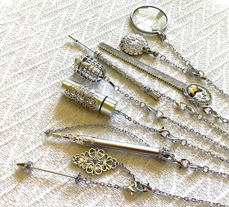 May include: A collection of silver-toned metal accessories including a magnifying glass, a ruler, a cylindrical container with filigree detail, a pen, and decorative charms, all connected by delicate silver chains with small clear crystal accents.