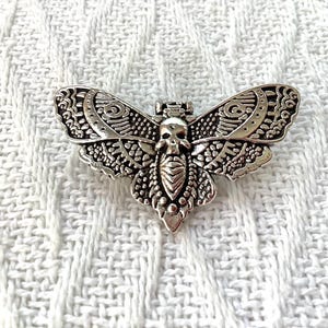 May include: A silver-toned metal brooch shaped like a moth with a skull design in the centre. The moth has intricate details and a textured surface.