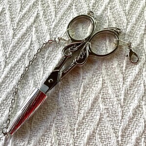 May include: Silver-toned embroidery scissors with ornate handles and a chain. The scissors have a cylindrical handle and a small clasp. The blades are closed. The scissors are on a white textured surface.
