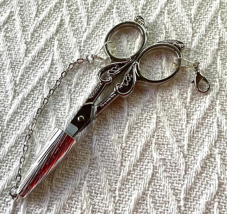 May include: Silver-toned embroidery scissors with ornate handles and a chain. The scissors have a cylindrical handle and a small clasp. The blades are closed. The scissors are on a white textured surface.