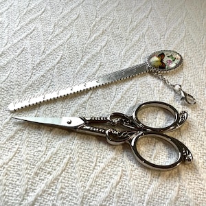 May include: A silver-toned metal bookmark featuring a butterfly graphic and a ruler marked in inches, paired with a pair of ornate silver-toned metal embroidery scissors with decorative scrollwork on the handles and sharp, pointed blades.